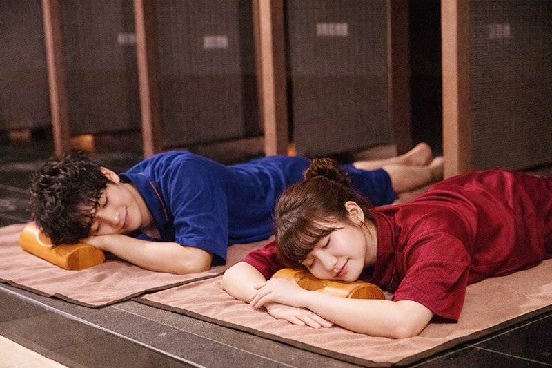 岩盤浴室内で、木製の枕に頭をのせてうつ伏せで横になる男女。